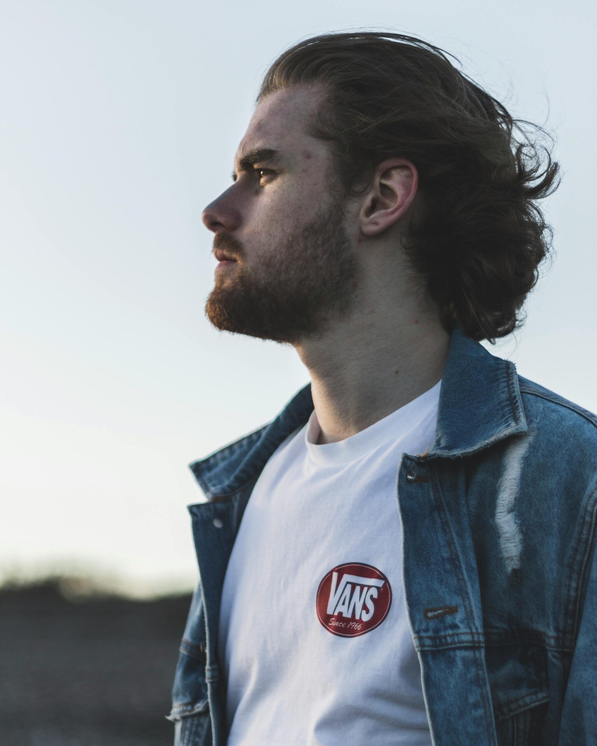 Profile of a fashionable man in a denim jacket outdoors, exuding a serious demeanor.